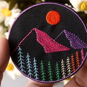 May include: Embroidered patch with a black background, featuring a pink and purple mountain range with a bright orange sun above. The mountains are surrounded by a row of colorful trees.