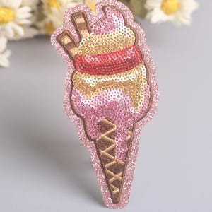 Peut inclure: Écusson de glace à la crème à paillettes avec des accents roses, dorés et rouges. Le cornet est marron et la glace a un tourbillon rose et doré avec une bande rouge. Deux gaufrettes brunes sont sur le dessus. L'écusson est bordé de paillettes.