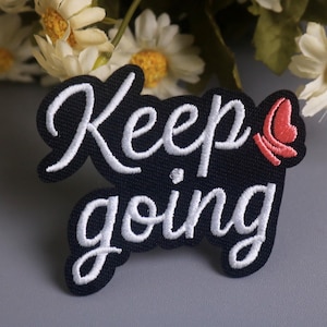 May include: A black embroidered patch with the words "Keep going" in white script. A small red butterfly is on the right side of the patch. The patch is on a gray surface with white flowers in the background.