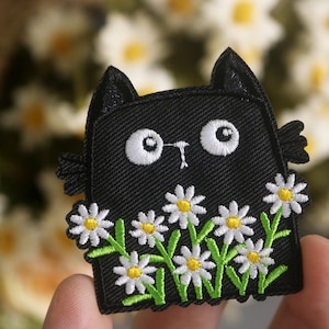 Peut inclure: Patch thermocollant brodé noir représentant un chat avec des yeux blancs et un nez blanc, entouré de marguerites blanches avec des centres jaunes et des tiges vertes.
