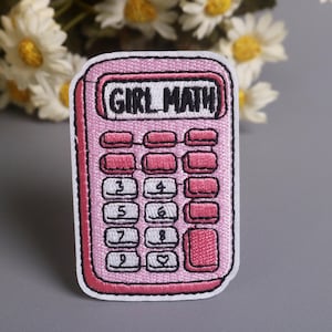 May include: Pink embroidered patch in the shape of a calculator with the words "GIRL MATH" in black. The calculator has pink buttons and a white border. Daisies are in the background.