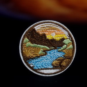May include: Circular embroidered patch depicting a landscape. The design shows a river winding through a valley, with mountains and a sunset. The patch has a white border, displaying a detailed nature scene.