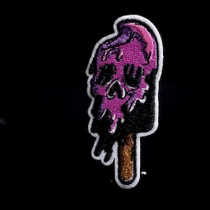 Op de afbeelding: Een paarse schedel-vormige ijslolly patch met een witte rand. De ijslolly smelt, met zwarte druppels. De stok is bruin. De patch staat op een zwarte achtergrond. De patch is een strijkapplicatie.