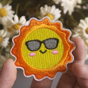 May include: A yellow embroidered sun with a smiling face wearing sunglasses. The sun is surrounded by an orange and white border.