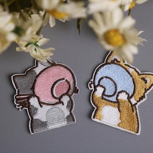 May include: Two embroidered patches featuring cartoon cats. One cat is gray, white, and pink, the other is brown, white, and blue. Both are holding a spiral object. The patches are on a gray background.