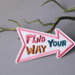 Op de afbeelding: Witte pijlvormige patch met roze stiksels. De patch heeft de tekst "FIND YOUR WAY" geborduurd in kleurrijk garen.