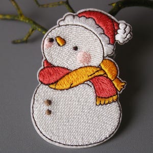 Puede incluir: Parche bordado para planchar de un muñeco de nieve con un sombrero rojo y una bufanda amarilla y roja. El muñeco de nieve tiene un cuerpo blanco, mejillas rojas y ojos negros.