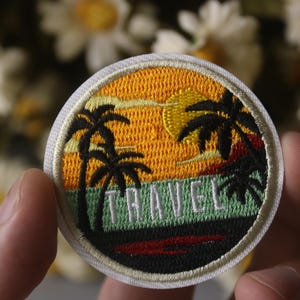 May include: A round embroidered patch with a sunset scene, palm trees, and the word "TRAVEL" in green text. The patch has a white border.