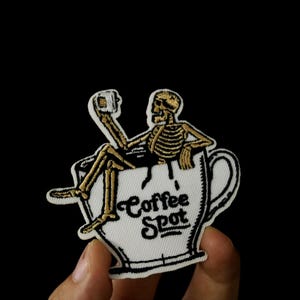 Op de afbeelding: Een witte koffiemok met een zwarte omtrek en de tekst "Coffee Spot" in zwarte letters. Een gouden skelet zit in de mok en houdt een kopje koffie vast.
