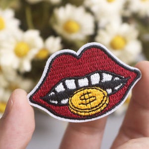 Könnte beinhalten: Gestickter Aufnäher mit roten Lippen mit weißen Zähnen, die eine Goldmünze mit einem Dollarzeichen halten. Der Aufnäher hat einen weißen Rand. Der Hintergrund zeigt verschwommene weiße und gelbe Blumen.