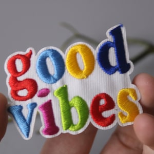 Puede incluir: Un parche bordado colorido con las palabras "good vibes" en colores arcoíris. El parche está hecho de tela blanca y tiene una superficie ligeramente texturizada.