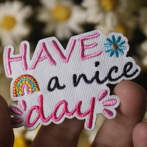 May include: White embroidered patch with pink lettering that says "Have a nice day". The patch also features a rainbow and a blue flower.