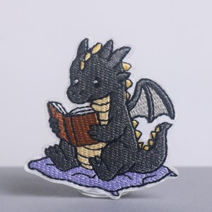 May include: Embroidered patch featuring a cartoon dragon reading a book. The dragon is black with yellow accents, sitting on a purple pillow. The book is brown. The patch is designed for application on clothing or accessories.