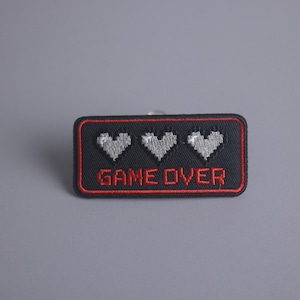 Op de afbeelding: Zwarte en rode geborduurde patch met drie witte gepixelde harten en de tekst "GAME OVER".