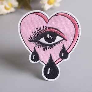May include: Embroidered patch in the shape of a pink heart with a black and white eye and black teardrops. The patch has a white border and is likely for clothing or accessories.