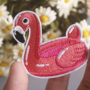 以下が含まれることがあります： 白いステッチが付いたピンクの浮き輪のフラミンゴのパッチ。パッチはピンクと白のデザインで刺繍されています。