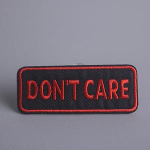Könnte beinhalten: Ein schwarzer rechteckiger Patch mit rotem gesticktem Text, der "DON'T CARE" lautet.