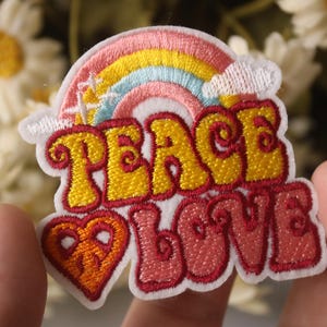 May include: Embroidered iron-on patch with a rainbow, peace sign, and the words "Peace Love" in a retro font. The patch is white with pink, yellow, orange, and blue embroidery.