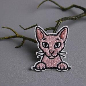 以下が含まれることがあります： 白い縁取りのピンク色の猫の刺繍パッチ。猫は大きな目、小さな鼻、ひげを持っています。パッチは、枝のある灰色の背景に配置されています。