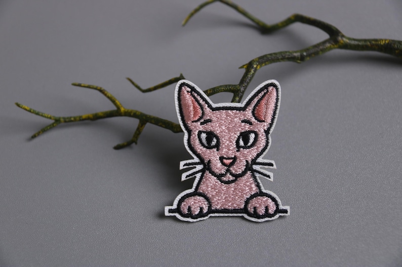 以下が含まれることがあります： 白い縁取りのピンク色の猫の刺繍パッチ。猫は大きな目、小さな鼻、ひげを持っています。パッチは、枝のある灰色の背景に配置されています。