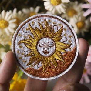 Könnte beinhalten: Ein runder gestickter Aufnäher mit weißem Hintergrund und einer goldenen Sonne mit einem lächelnden Gesicht. Die Sonne hat einen braunen und orangenen Hintergrund.