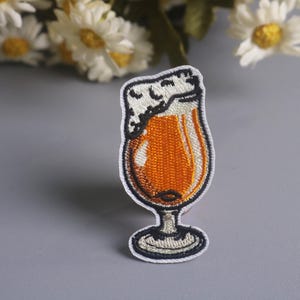 Op de afbeelding: Geborduurde patch met een bierglas gevuld met amberkleurige vloeistof en bedekt met wit schuim. De patch heeft een gedetailleerd ontwerp met zwart-witte stiksels die het glas en het schuim omlijnen. Voor kleding of accessoires.