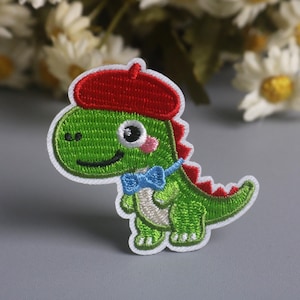 May include: Embroidered patch of a green dinosaur wearing a red beret and a blue bow tie. The dinosaur has a white outline and a smiling face. The patch is on a gray surface with white flowers in the background.