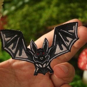 May include: Black and white embroidered iron-on patch of a bat with red eyes. The bat has a detailed design with wings and a sharp face.