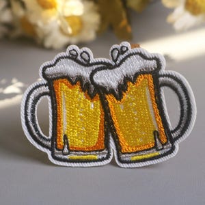 Può includere: Toppa ricamata con due boccali di birra che si toccano. I boccali sono pieni di liquido giallo, sormontati da schiuma bianca e bordati di nero e bianco. Per abbigliamento o accessori.