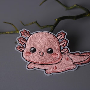 May include: Pink embroidered axolotl patch with a cute cartoon face and a white background.