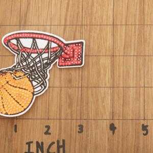 Peut inclure: Un &eacute;cusson de basket-ball repr&eacute;sentant un ballon de basket-ball traversant un filet. Le ballon est orange avec des lignes noires, et le filet est blanc et noir. Le cercle est rouge avec des points blancs. L'&eacute;cusson mesure environ 5 cm de large.
