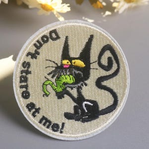 Op de afbeelding: Ronde geborduurde patch met een zwarte kat met gele ogen en een groen object in zijn mond. De tekst "Don't stare at me!" staat in een cirkel rond de kat.