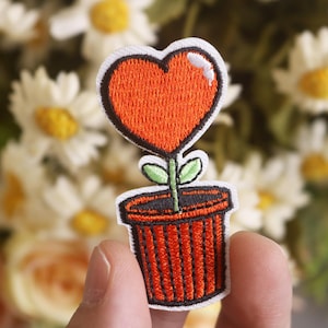 Puede incluir: Una flor roja en forma de corazón que crece en una maceta roja con hojas verdes. La flor y la maceta están bordadas en una tela blanca.