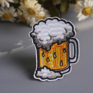 May include: Embroidered patch depicting a beer mug with a foamy head. The mug is yellow with vertical lines, and the foam is white and grey. The patch has a black border and is suitable for clothing or accessories.