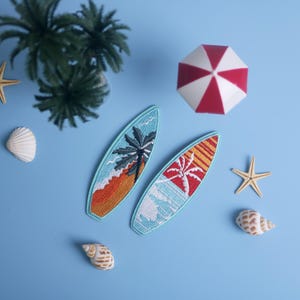 Op de afbeelding: Twee geborduurde surfplankpatches met palmboomontwerpen, &eacute;&eacute;n met oranje en de andere met rode accenten, worden weergegeven op een lichtblauwe ondergrond. De sc&egrave;ne bevat een rood-witte paraplu, zeesterren, schelpen en een palmboom.