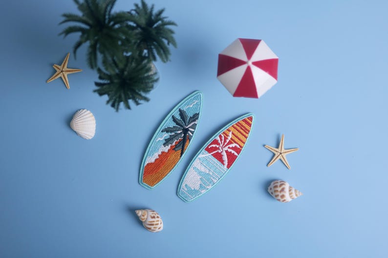 Op de afbeelding: Twee geborduurde surfplankpatches met palmboomontwerpen, &eacute;&eacute;n met oranje en de andere met rode accenten, worden weergegeven op een lichtblauwe ondergrond. De sc&egrave;ne bevat een rood-witte paraplu, zeesterren, schelpen en een palmboom.