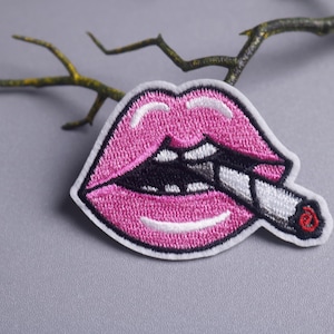 Könnte beinhalten: Gestickter Aufnäher mit einem rosa Mund mit einer Zigarette. Die Lippen sind leuchtend rosa mit weißen Details, und die Zigarette ist weiß mit einer roten Spitze. Der Aufnäher hat einen weißen Rand.