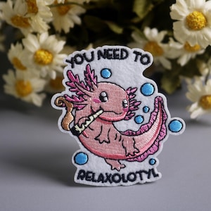Puede incluir: Parche bordado con un ajolote rosa fumando un porro, rodeado de burbujas azules. El texto "YOU NEED TO RELAXOLOTL" está arriba y abajo del ajolote. El parche está sobre un fondo blanco.