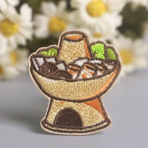 May include: Embroidered patch featuring a cartoon depiction of a hot pot. The patch has a tan and brown color scheme, with green and brown food items inside the pot. The patch is on a gray surface with white flowers in the background.
