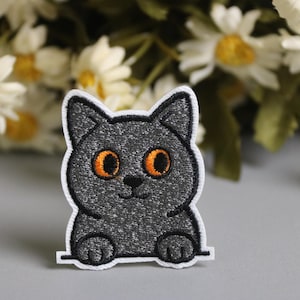 May include: A gray cat patch with orange eyes and a white border. The embroidered patch features a smiling cat with its paws resting on a ledge. The background includes white daisies with yellow centers.