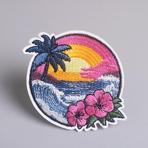 Op de afbeelding: Geborduurde patch met een tropische zonsondergang met een palmboom, oceaangolven en roze hibiscusbloemen. Het ontwerp is geplaatst in een cirkelvormige rand, met levendige kleuren blauw, geel, roze en groen.