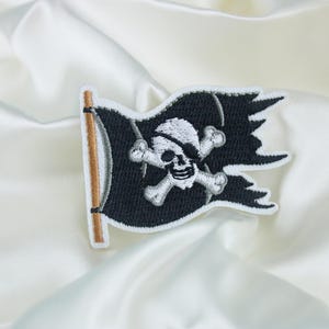 Può includere: Toppa di bandiera pirata ricamata in bianco e nero con un design di teschio e ossa incrociate. La toppa è su uno sfondo di tessuto bianco.