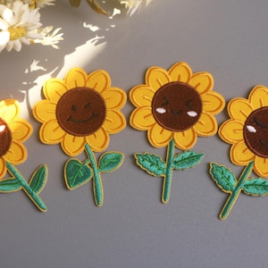 May include: Four embroidered sunflower patches with smiling faces. Each patch features yellow petals, a brown centre, green leaves, and a green stem. The patches are arranged on a grey surface.