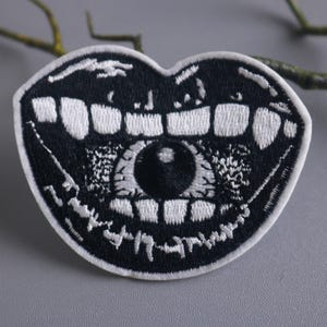 May include: Black and white embroidered patch depicting a large open mouth with a single eye in the centre. The mouth has sharp teeth and the eye is staring directly at the viewer.