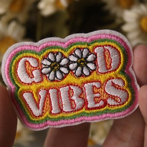Puede incluir: Un parche bordado de color rosa, verde y amarillo con las palabras "GOOD VIBES" en blanco. Dos margaritas blancas con centros amarillos están bordadas encima de las palabras.