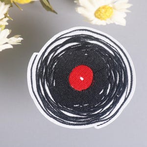 Puede incluir: Parche bordado con un diseño de disco de vinilo negro con un centro rojo. El parche tiene un borde blanco y es adecuado para su aplicación en ropa o accesorios. El diseño es una representación estilizada de un disco.
