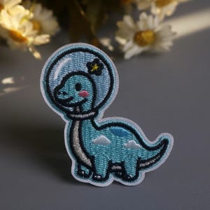 May include: Embroidered patch featuring a cartoon dinosaur in a space helmet. The dinosaur is light blue with a smiling face, wearing a clear helmet with a yellow flower. The patch has a white border and is suitable for clothing or accessories.