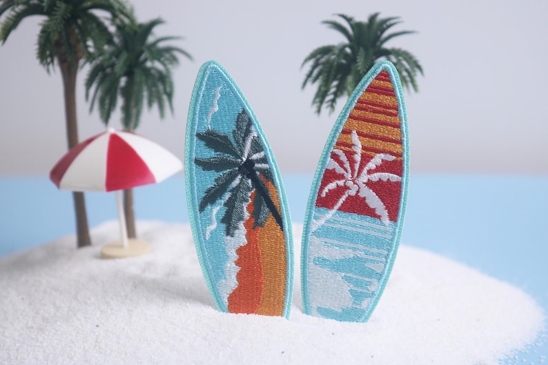 Op de afbeelding: Twee geborduurde surfplankpatches met tropische strandsc&egrave;nes. E&eacute;n heeft een palmboom en een oranje zonsondergang, de andere een palmboom en een rode zonsondergang. Ze staan op een zanderig eiland met palmbomen en een rood-witte parasol.