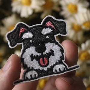 May include: An embroidered patch of a smiling schnauzer dog, with black and white fur and a pink tongue. The patch is held in a hand against a blurred background of white flowers.