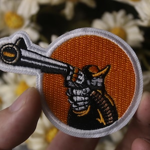 May include: An embroidered patch featuring a cartoon cowboy aiming a gun. The cowboy is wearing a black hat and a brown vest. The background is orange.